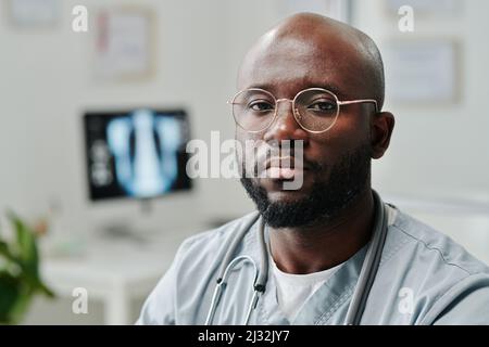 Junger ernsthafter afroamerikanischer Hausarzt in blauer Uniform und Brillen, der am Arbeitsplatz vor der Kamera im Krankenhaus sitzt Stockfoto