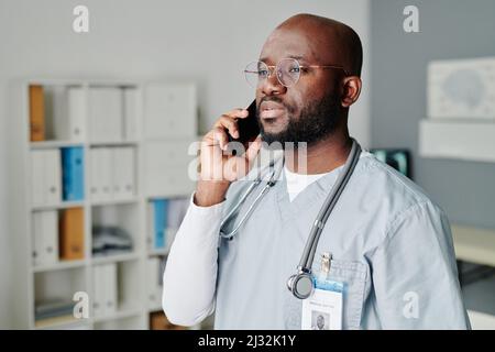 Selbstbewusster männlicher Arzt mit Stethoskop am Hals, der mit dem Patienten auf dem Mobiltelefon spricht, während er im medizinischen Büro vor der Kamera steht Stockfoto