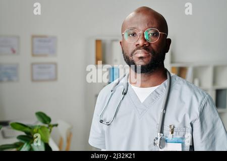 Junger, zeitgenössischer afroamerikanischer Klinikarzt in Brillen und blauer Uniform, der im medizinischen Büro steht und die Kamera anschaut Stockfoto