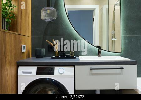 Duftkerze, Reed-Lufterfrischer und schwarze Glas-Nachfüllflasche auf der Waschmaschine neben dem Waschbecken mit Lampe und halbkreisförmigen Spiegel darüber Stockfoto