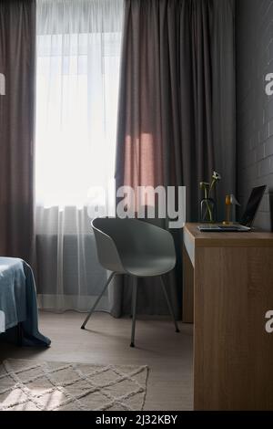 Ecke des Wohnzimmers mit Laptop auf Holzschreibtisch und grauem Plastiksessel, der gegen das Fenster mit weißen und grauen Vorhängen steht Stockfoto