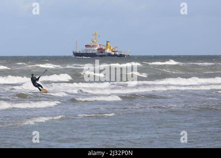 05. April 2022, Mecklenburg-Vorpommern, Warnemünde: Ein Kitesurfer gleitet bei starkem Wind durch die Ostsee, während ein Forschungsschiff den Eingang von Warnemünde verlässt. Der Wasserstand der Ostsee vor Warnemünde hat sich nach der Ebbe der letzten Tage wieder normalisiert. Foto: Danny Gohlke/dpa Stockfoto