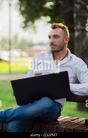 Verträumter Mann arbeitet im Park mit einem Laptop, trinkt Kaffee. Ein junger Mann auf einem Hintergrund von grünen Bäumen, ein heißer sonniger Sommertag. Warmes, weiches Licht, Nahaufnahme. Stockfoto