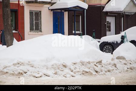 Eine lockere Schneeverwehung an der Straße vor dem Hintergrund einer Stadtstraße mit Autos. Auf der Straße liegt schmutziger Schnee in hohen Haufen. Urbane Winterlandschaft. Wolkiger Wintertag, weiches Licht. Stockfoto