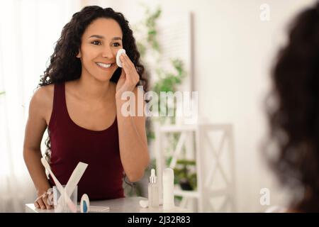 Schöne junge Frau, die Make-up mit Wattepad entfernt, Hauttonikum aufgetragen, Spiegel angeschaut, im Badezimmer Platz kopiert Stockfoto