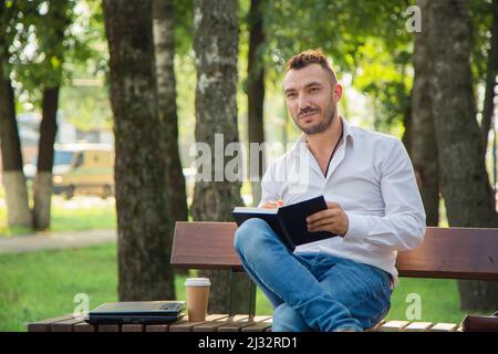 Ein lächelnder Mann sitzt auf einer Bank, macht Pläne, schreibt in ein Notizbuch. Ein junger Mann auf einem Hintergrund von grünen Bäumen, ein heißer sonniger Sommertag. Warmes, weiches Licht, Nahaufnahme. Stockfoto