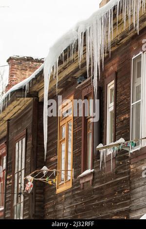 Scharfe transparente Eiszapfen hängen am Rand des Dachs. Vor dem Hintergrund der Holzwand des alten Hauses. Große Kaskaden, sogar schöne Reihen. Wolkiger Wintertag, weiches Licht. Stockfoto