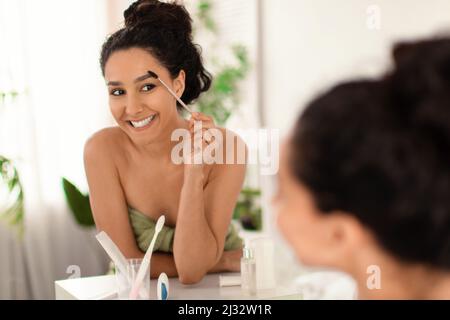 Schöne junge Frau, die Mascara anwendet, im Spiegel auf das Badezimmer schaut, leerer Raum Stockfoto