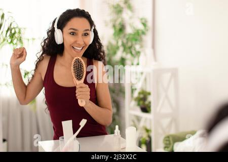 Fröhliche junge Brünette weiblich in Kopfhörern tanzen und singen, mit Haarbürste als Mikrofon in der Nähe des Spiegels zu Hause Stockfoto