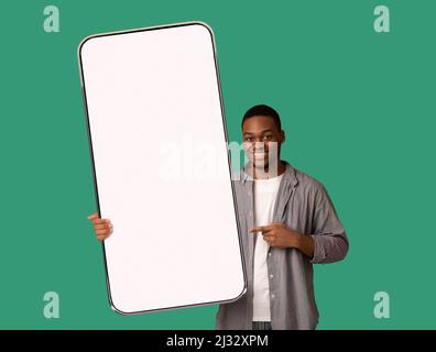 Tolles Angebot für Mobilgeräte. Glücklicher schwarzer Mann zeigt auf riesige leere Smartphone in der Hand, zeigt Modell für Ihr Design Stockfoto