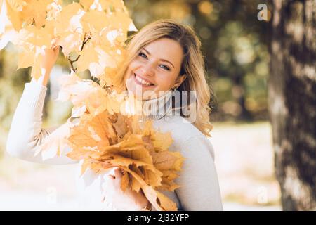 Lachende blonde Frau, die ein paar goldene Blätter mit Ahorn in zwei Händen neben dem Baum hält, verschönert die Unschärfe. Schließen Stockfoto