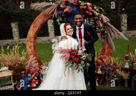 Schöne weiße Frau mit charmanten afroamerikanischen Mann stehen in der Nähe dekorierten Bogen, entzückendes Paar auf Hochzeitszeremonie, attraktive Bräutigam Umarmung schöne b Stockfoto