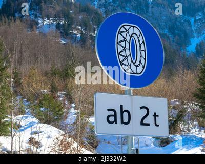 Nahaufnahme eines europäischen Schildes, das darauf hinweist, dass im Winter Schneekettenräder im Eis fahren müssen. Winterlandschaft und Wald im Hintergrund. Stockfoto