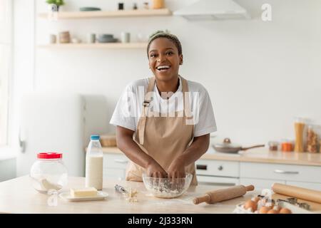 Lachende junge afroamerikanische Dame auf der Schürze, die in minimalistischer Küchenausstattung Pie-Teig machte Stockfoto