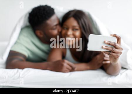 Lächelnder, tausendjähriger afroamerikanischer Mann, der Frau küsst, Frau, die Selfie auf dem Smartphone auf dem weißen Bett macht Stockfoto