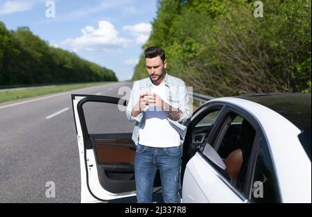 Verärgert junger Mann, der in der Nähe des Autos steht, Smartphone anschaut, Probleme mit der mobilen Verbindung hat und versucht, ein Netzwerk zu finden Stockfoto