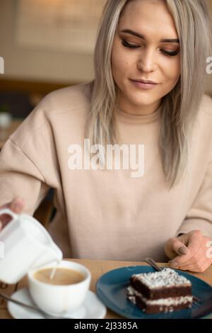 Porträt einer jungen, herrlich lächelnden Frau mit langen grauen Haaren mit perfektem Make-up, das Milch in eine Tasse Kaffee gießt. Stockfoto