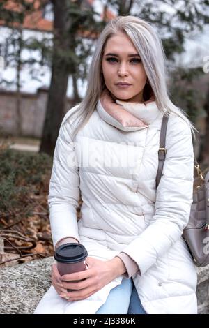 Porträt einer jungen schönen ernsthaften Frau mit langen Haaren mit Make-up in weißer Jacke mit einer Tasse Kaffee zum Mitnehmen. Stockfoto