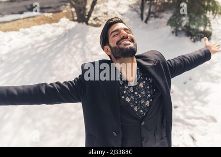 Stilvoller, gepflegter Mann aus Latein, der den sonnigen Wintertag mit Schnee genießt - in schwarzem Mantel und Anzug gekleidet. Hochwertige Fotos Stockfoto