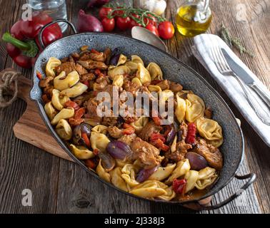 Gebratenes Schweinefleisch in der Pfanne mit Gemüse, Sauce und Tortellini in einer Bratpfanne auf einem Holztisch Stockfoto