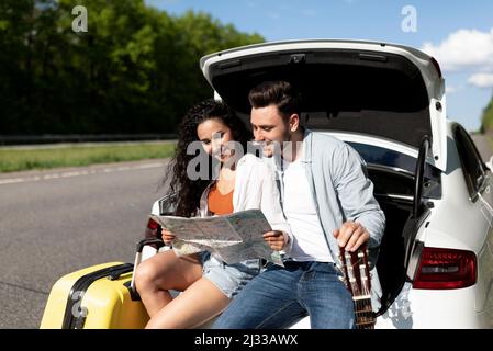 Positives Paar, das im Auto unterwegs ist, auf der Karte nachschaut, Pause vom Fahren macht, zusammen im Auto-Kofferraum sitzt Stockfoto