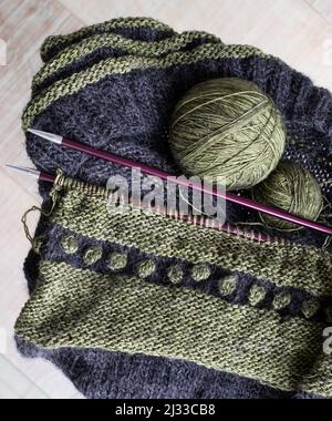 Grüner und grauer Wollpullover wird gerade gestrickt. Das Foto zeigt strukturiertes Stricken inklusive Strumpfstich, Rippe, Noppen, Streifen. Nadeln. Stockfoto