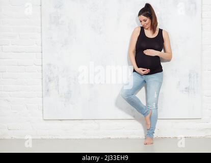 Modetrorträt der glücklichen Schwangeren in Jeans. Gegen weiße Wand mit Copyspace. Stockfoto