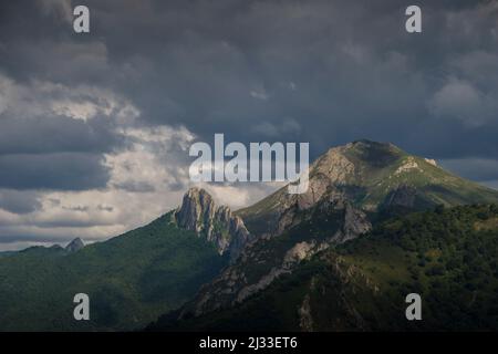 Atemberaubende Sonnenstrahlen leuchten durch einen bewölkten Himmel auf kantigen Berggipfeln über grünem Wald Stockfoto