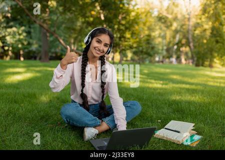 Ich mag E-Learning. Glückliche indische Studentin mit Laptop, mit Online-Klasse und zeigen Daumen nach oben, sitzen im Freien Stockfoto