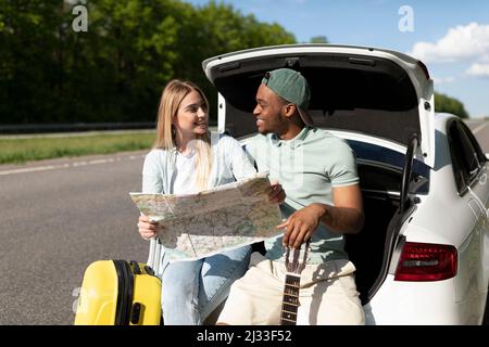 Liebevoller schwarzer Kerl und seine Freundin studieren Karte, während sie auf ihren Reisen im Kofferraum sitzen Stockfoto