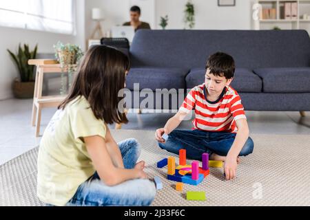 Zwei Kinder spielen zusammen auf dem Boden, während ihr Vater mit einem Laptop im Hintergrund arbeitet. Stockfoto