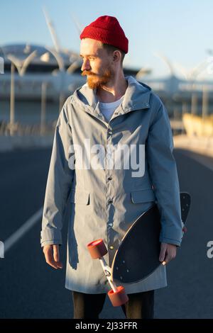 Lässiger Mann mit Longboard über dem Stadtgebäude. Stylischer Hipster-Skateboarder hält Skateboard bei Sonnenuntergang Stockfoto