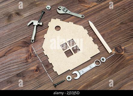 Unverzichtbare Handwerkzeuge und Befestigungselemente, die um eine Miniatur-flache Hausform auf einem Holzhintergrund angeordnet sind. Haushaltsverbesserungs- und Reparaturkonzept. Stockfoto