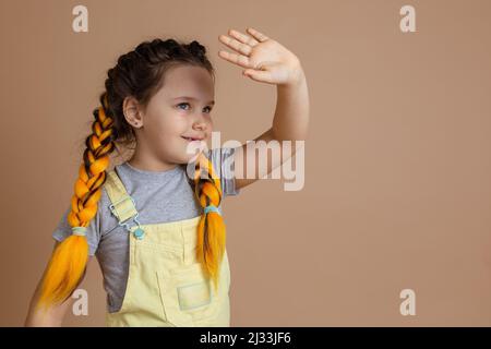 Kleines glückseliges kaukasisches Mädchen mit gelben Kanekalon-Zöpfen, das mit lächelnder Hand auf jemanden verzichtet und in gelbem Jumpsuit und grauem T-Shirt wegschaut Stockfoto