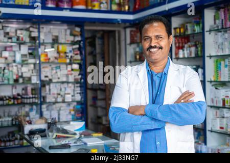 Glücklicher Apotheker in Uniform stehend mit gekreuzten Armen durch Blick auf Kamera im Einzelhandel medizinischen Laden oder Shop mit Kopieplatz - Konzept der kleinen Stockfoto