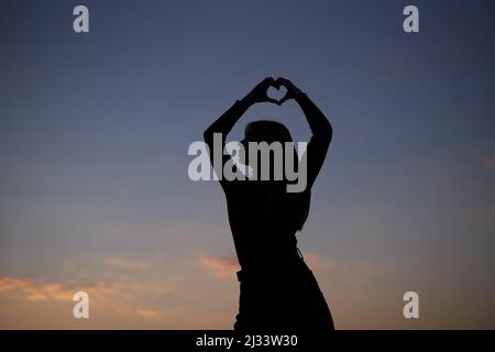Attraktive kaukasische Frau mit langen Haaren, die vor der Kamera in Silhouette posiert und Liebe oder Herzensgestik im Freien bei Sonnenuntergang mit Blick auf den Himmel zeigt. Romantik- oder Liebeskonzept. Hochwertige Bilder Stockfoto