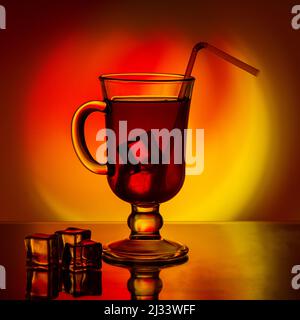 Kalter Tee, Cocktail, Fruchtgetränk in einer hohen Glastasse mit Trinkhalm auf einem Tisch mit Eisstücken auf einem schönen, leuchtend gelb-roten Farbverlauf zurück Stockfoto