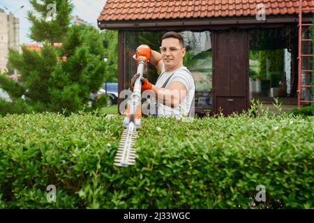 Hübscher junger Mann in Schutzbrille und Handschuhen, der grüne Büsche auf dem Hinterhof formt. Professioneller männlicher Gärtner, der eine Benzinschneidmaschine für die Pflege von Pflanzen verwendet. Stockfoto