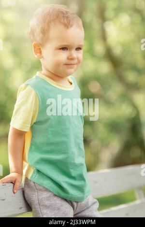 Wohlmeinend, glücklicher Junge ruht im Garten, er beobachtet ruhig die Umgebung und lächelt. Sonniger Sommertag, fröhliches Kind mit grünem Hemd, gelbe s Stockfoto