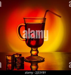 Kalter Tee, Cocktail, Fruchtgetränk in einer hohen Glastasse mit Trinkhalm auf einem Tisch mit Eisstücken auf einem schönen, leuchtend gelb-roten Farbverlauf zurück Stockfoto