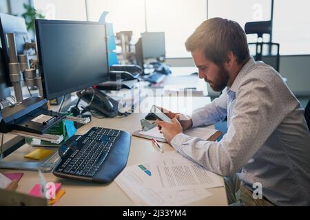 Lesen einer wichtigen Botschaft. Aufnahme eines fokussierten jungen Geschäftsmannes, der auf seinem Handy SMS schreibt, während er hinter seinem Schreibtisch im Büro sitzt. Stockfoto