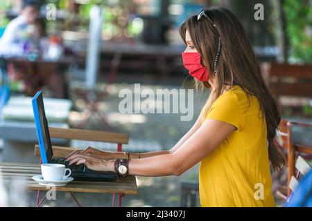 Frau sitzt in leerem Café mit Schutzmaske auf Laptop arbeiten Stockfoto