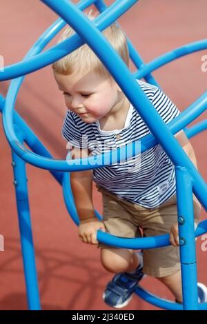 Fokussierter Junge klettert auf die blaue, rohrförmige Spielplatzleiter. Frech Kind genießen sonnigen Tag in einer Öffentlichkeit. Kümmert sich nicht um das Risiko Stockfoto