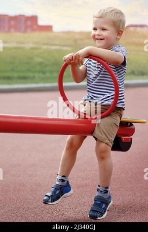 Schöner Junge sitzt auf einer doppelseitigen Schaukel auf einem Spielplatz. Lächelnder Junge im Sommer. Gerne erhöht werden. Fröhliches Kind draußen Stockfoto