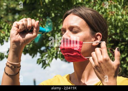 Junge Frau kühlt sich mit einem Handventilator ab, während sie eine Gesichtsschutzmaske trägt Stockfoto