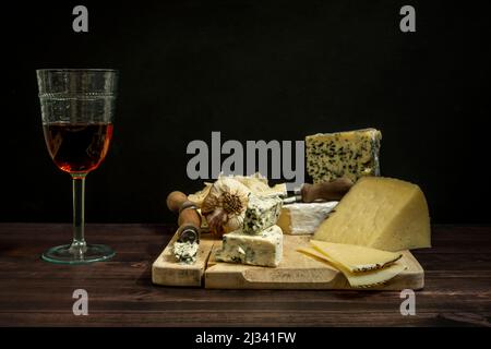 Stillleben mit Manchego gehäutetem Schafskäse und französischem Blaukäse mit Messern und Knoblauch und einem Glas spanischem Roséwein Stockfoto