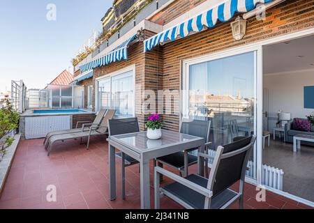 Penthouse-Terrasse mit blauen und weißen Markisen, grauem Metall- und Glastisch, passenden Stühlen, Sonnenliegen und einem kleinen Spa-Pool Stockfoto