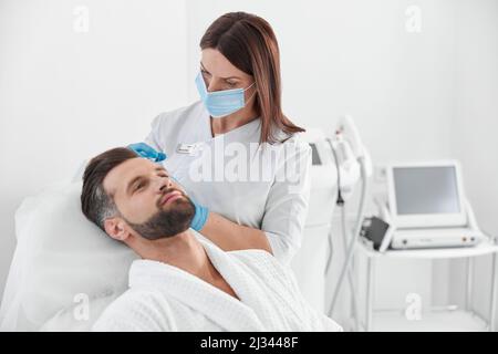 Die Kosmetologin mit Maske spritzt bei der Meso-Therapie in die Kopfhaut des Patienten Stockfoto