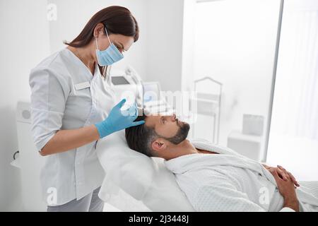 Professionelle Kosmetikerin hält Spritze mit Füllstoff über der Stirn des Kunden in der Kosmetologie-Klinik Stockfoto