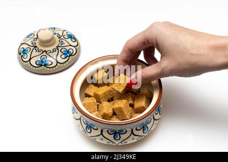 Die Hand einer Frau nimmt einen Würfel braunen Zuckers aus einer Zuckerschüssel Stockfoto